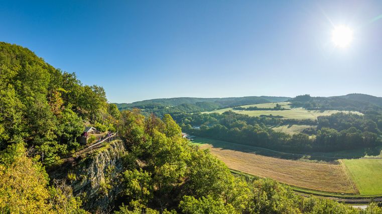 Eine malerische Landschaft mit sanften Hügeln und einem klaren blauen Himmel. Im Vordergrund sind grüne Bäume und eine Wiese zu sehen.
