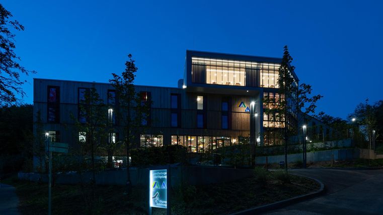 Ein modernes Gebäude bei Nacht, beleuchtet und umgeben von Bäumen. Die Fenster strahlen Wärme aus und schaffen eine einladende Atmosphäre.