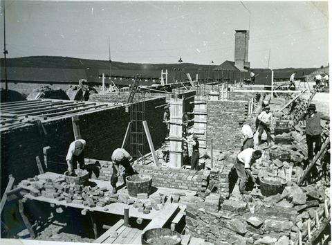 Eine Baustelle mit mehreren Arbeitern, die Steine verlegen. Im Hintergrund sind Gebäudestrukturen und eine Industrieanlage zu sehen.