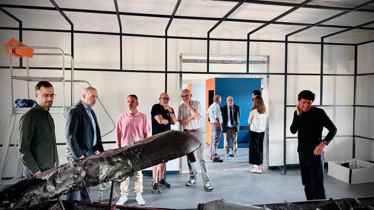 Eine Gruppe von Menschen steht in einem modernen Raum mit einer künstlerischen Installation. Die Atmosphäre ist gespannt, während die Besucher die Exponate betrachten.