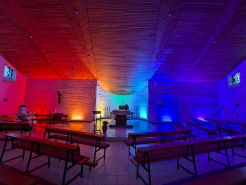 Eine moderne Kirche mit bunten Lichtern in Rot, Blau und Violett. Bänke sind in einem offenen Raum angeordnet, der eine ruhige Atmosphäre ausstrahlt.