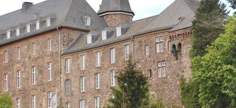Un bâtiment historique avec un toit gris et une façade en pierre. Entouré d'arbres verts et d'un paysage pittoresque.