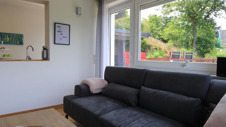 Ein modernes Wohnzimmer mit einem grauen Sofa und großen Fenstern. Draußen ist eine grüne Landschaft mit Bäumen und Bänken zu sehen.