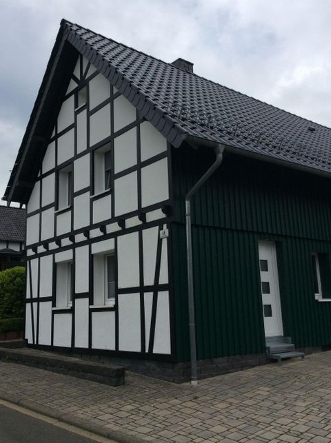 Ein modernes Fachwerkhaus mit einer Kombination aus grüner und weißer Fassade. Das Dach ist schwarz und der Himmel ist bewölkt.