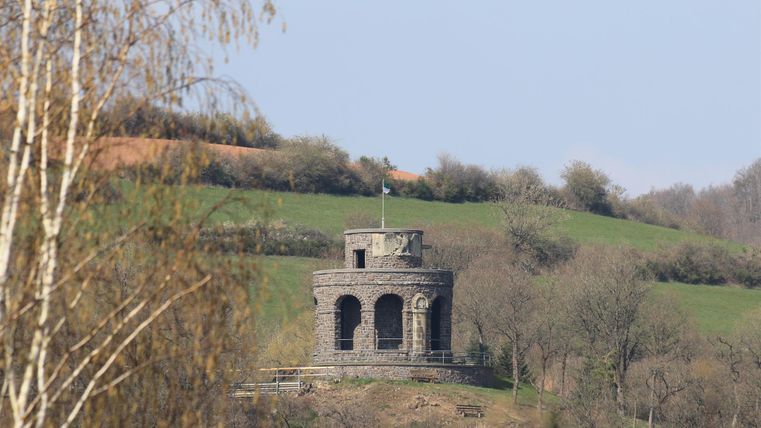Ein historisches Denkmal aus Stein steht auf einem bewaldeten Hügel. Im Hintergrund sind sanfte grüne Landschaften und ein blauer Himmel zu sehen.