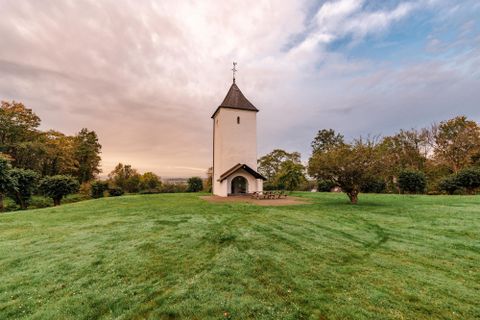 Eine kleine Kirche steht auf einer grünen Wiese. Im Hintergrund sind Bäume und ein sanfter Himmel zu sehen.