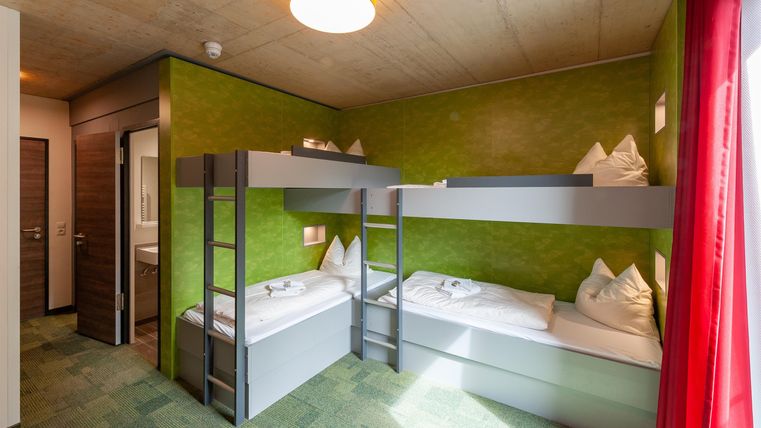 Ein farbenfrohes Schlafraum mit Etagenbetten und einem Einzelbett. Die Wände sind grün gestrichen, und große Fenster lassen viel Licht herein.