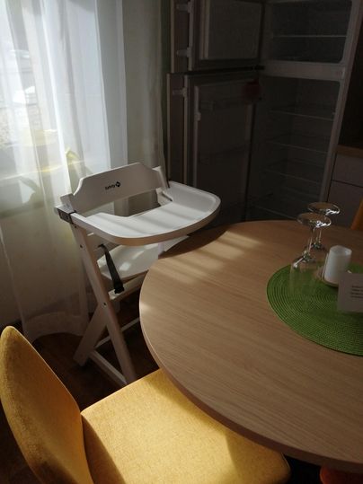 Ein Hochstuhl steht in einer hellen Küche neben einem runden Tisch. Der Raum ist gut beleuchtet, mit gelben Stühlen und einem grünen Tischset.