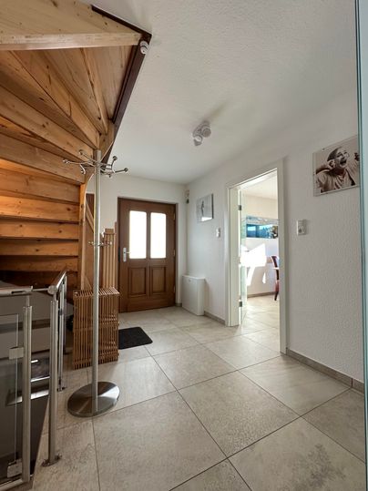 Ein heller Flur mit Treppe aus Holz und einem Eingangstür. Rechts ist ein weiterer Raum sichtbar, der gut beleuchtet ist.