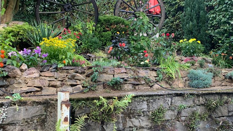 Ein blühender Garten mit bunten Blumen und einer Steineinfassung. Im Hintergrund sind zwei alte Waggonräder und ein roter Sonnenschirm zu sehen.