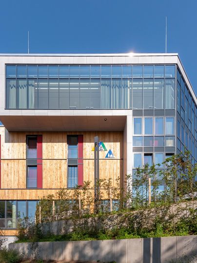 Ein modernes Gebäude mit großen Glasfenstern und holzverkleideter Fassade. Der Himmel ist klar und blau, umgeben von grünen Pflanzen.