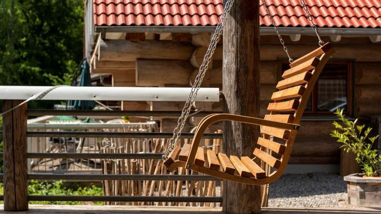 Eine hängende Holzschaukel an einem Holzhaus. Im Hintergrund sind Gartenmöbel und Pflanzen zu sehen.