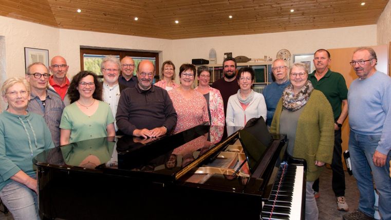 Eine Gruppe von Menschen steht um ein Klavier in einem gemütlichen Raum. Sie lächeln alle freundlich und scheinen eine gesellige Zeit zu haben.