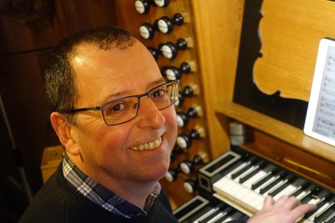 Ein Mann sitzt an einer Orgel und lächelt in die Kamera. Im Hintergrund sind die Orgelpfeifen und Notenblätter sichtbar.