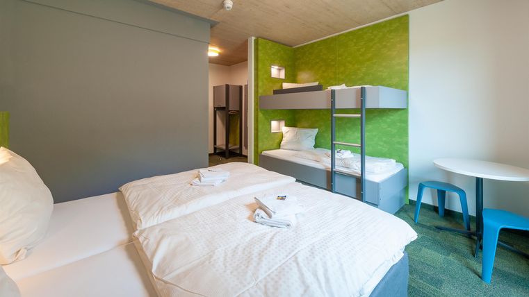 Ein modernes Hotelzimmer mit zwei Betten und einem Etagenbett. Die Wände sind in frischen Farben gestaltet, und es gibt einen kleinen Tisch mit Stühlen.