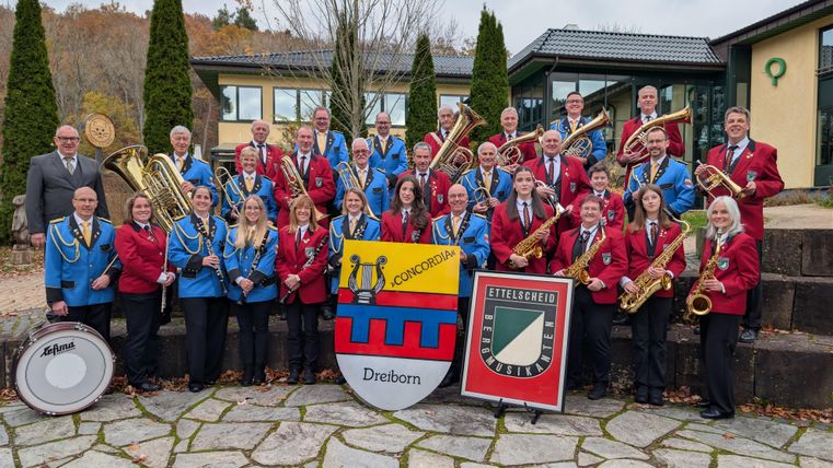 Eine große Blaskapelle steht im Freien und posiert für ein Gruppenfoto. Die Musiker tragen farbige Uniformen und halten ihre Instrumente.