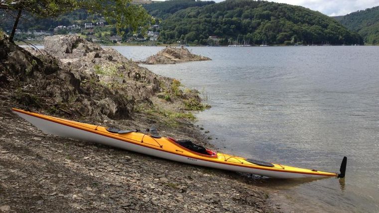 Ein Kajak liegt am Ufer eines ruhigen Gewässers. Sanfte Hügel und Bäume säumen den Hintergrund.