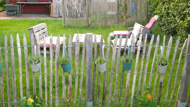 Ein Garten mit einem Holzzaun, an dem Blumenpflanzen hängen. Im Hintergrund stehen zwei Gartenstühle und ein Tisch.