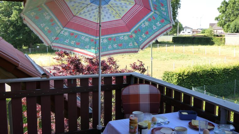 Ein Balkon mit einem bunten Sonnenschirm und einem gedeckten Tisch für das Frühstück. Im Hintergrund ist eine grüne Wiese zu sehen.