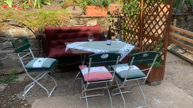 Ein gemütlicher Sitzbereich im Garten mit einer Bank und einem Tisch. Um den Tisch stehen vier Klappstühle mit karierten Kissen.