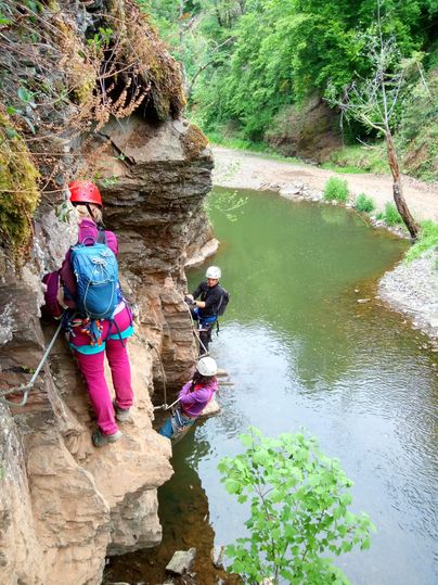 Eine Gruppe von Personen klettert an einer Felswand entlang eines kleinen Flusses. Die Umgebung ist grün und von Bäumen umgeben.