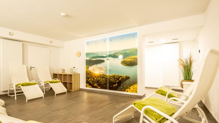 Ein heller Entspannungsraum mit Liegen und einer schönen Landschaftsandeutung an der Wand. Grünes Zubehör sorgt für eine einladende Atmosphäre.