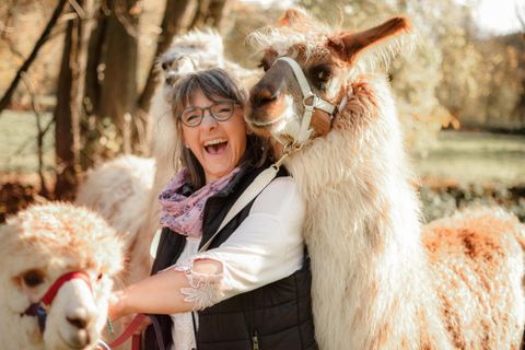 Een blije vrouw omarmt een lama en glimlacht naar de camera. Op de achtergrond zijn er meer lama's en herfstige bomen te zien.