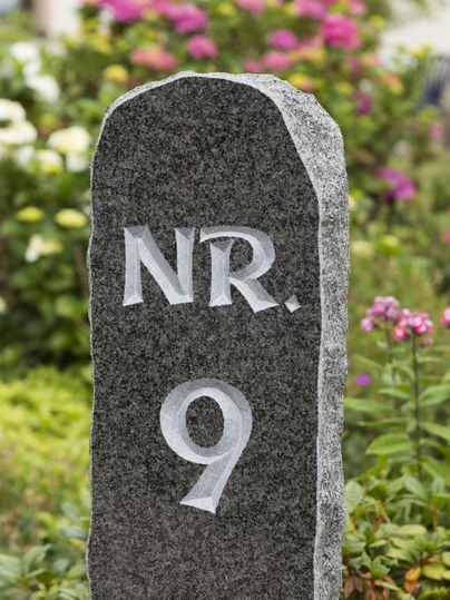 Ein schwarzer Steinsockel mit der Nummer 9. Im Hintergrund blühen bunte Blumen.
