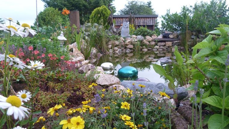 Ein schöner Garten mit bunten Blumen und einem kleinen Teich. Im Hintergrund sind Steine und Grünpflanzen zu sehen.
