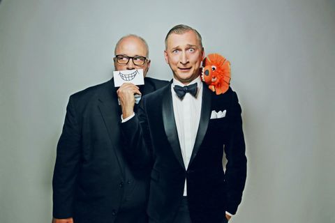 Zwei Männer in formeller Kleidung stehen zusammen. Der Mann vorne hält ein großes Lächeln-Schild und hat einen orangefarbenen Plüschkopf auf seiner Schulter.