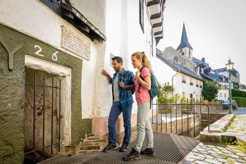 Ein Paar steht vor einem alten, historischen Gebäude mit einer kleinen Tür. Im Hintergrund sind malerische Fachwerkhäuser und ein Kirchturm zu sehen.