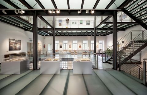 Ein modernes Kunstmuseum mit offenem Raum und großen Fenstern. Im Vordergrund stehen weiße Ausstellungsplattformen und im Hintergrund sind Bilder an den Wänden zu sehen.
