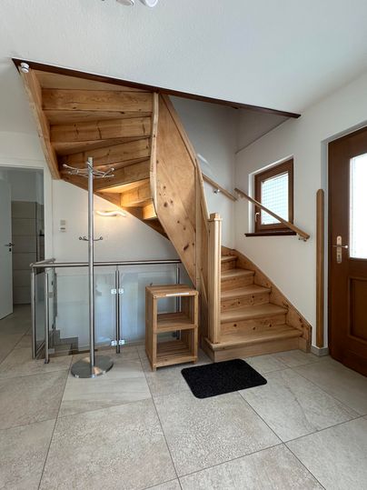 Ein heller Flur mit einer Holztreppe und großen Fenstern. Der Boden ist mit hellen Fliesen ausgelegt, und eine kleine Garderobe steht im Raum.