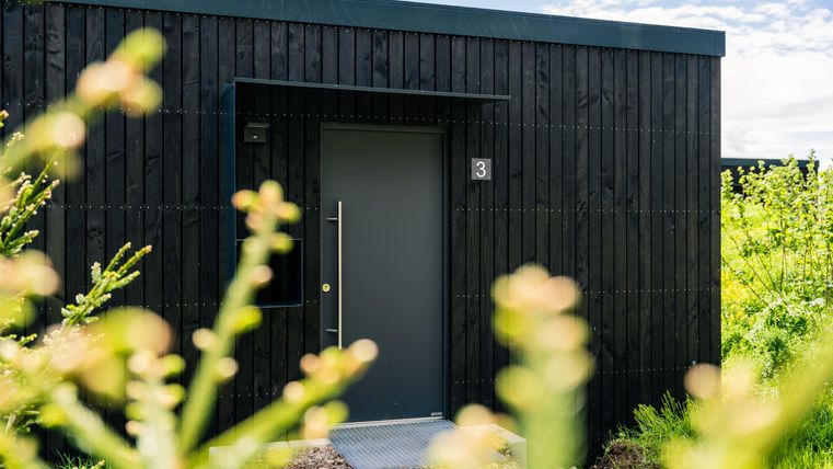 Ein modernes schwarzes Gebäude mit einer hellen Tür. Im Vordergrund sind grüne Pflanzen sichtbar.