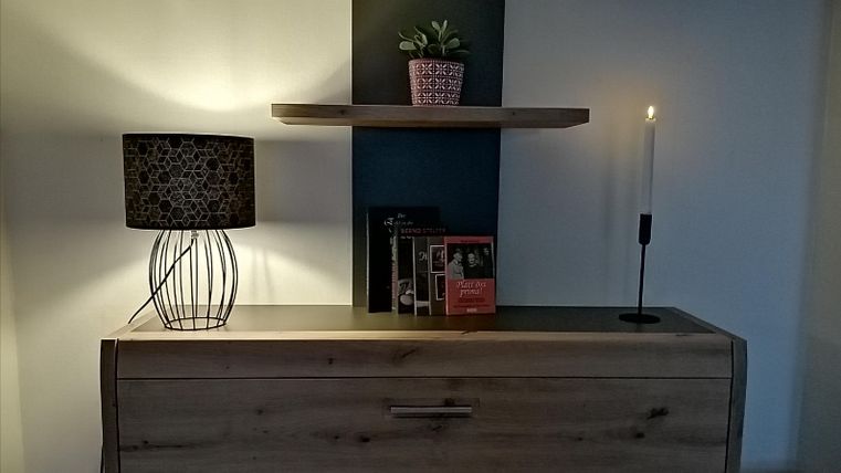 Ein modernes Sideboard aus Holz mit einer Lampe und einem Kerzenhalter. Auf dem oberen Regal steht eine kleine Pflanze und einige Bücher.