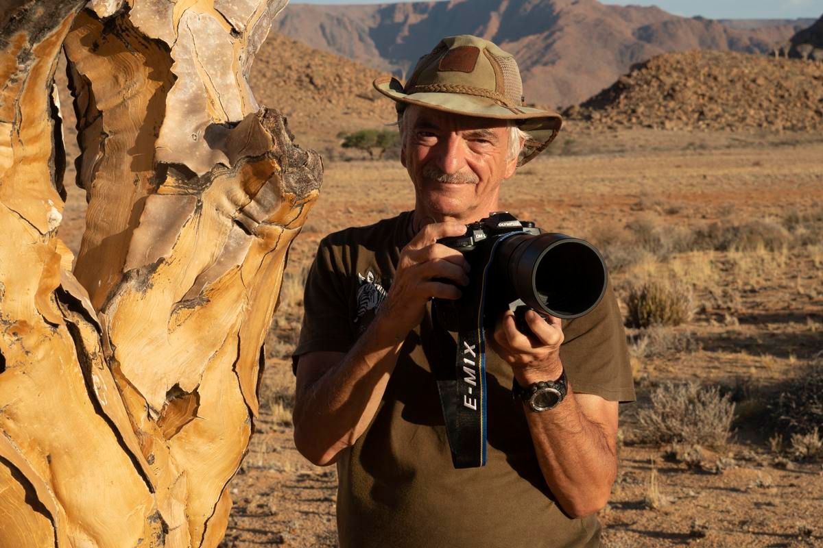 Ein älterer Mann mit einem Hut hält eine Kamera in der Hand. Im Hintergrund ist eine trockene, felsige Landschaft zu sehen.
