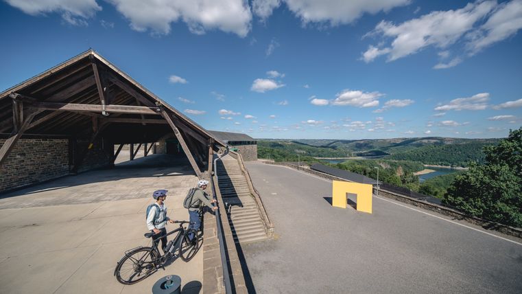 Zwei Radfahrer stehen vor einem Gebäude mit Holzdach und blicken auf eine hügelige Landschaft mit Fluss.