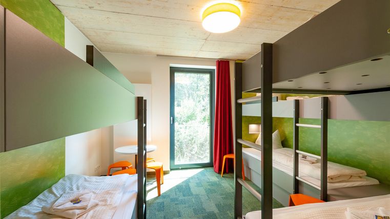 Ein modernes Zimmer mit Etagenbetten und bunten Möbeln. Große Fenster sorgen für viel Tageslicht.