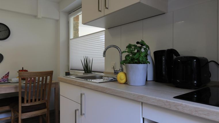 Eine moderne Küche mit hellen Möbeln und einer kleinen Pflanze auf der Arbeitsplatte. Im Hintergrund steht ein Holzstuhl und ein Tisch mit Zubehör.