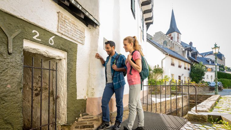 Ein Paar steht vor einem alten, historischen Gebäude mit einer kleinen Tür. Im Hintergrund sind malerische Fachwerkhäuser und ein Kirchturm zu sehen.