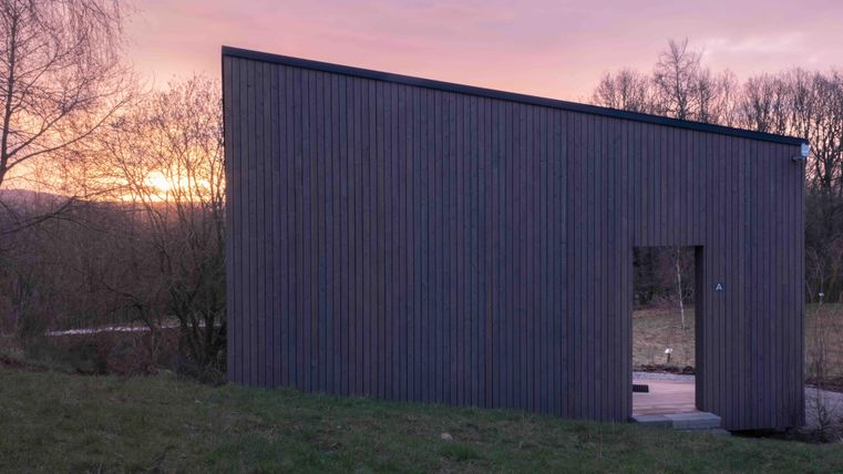 Ein modernes Holzhaus mit schräg verlaufendem Dach steht auf einer Wiese. Im Hintergrund ist ein farbenfroher Sonnenuntergang zu sehen.