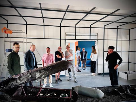 Eine Gruppe von Menschen steht in einem modernen Raum mit einer künstlerischen Installation. Die Atmosphäre ist gespannt, während die Besucher die Exponate betrachten.