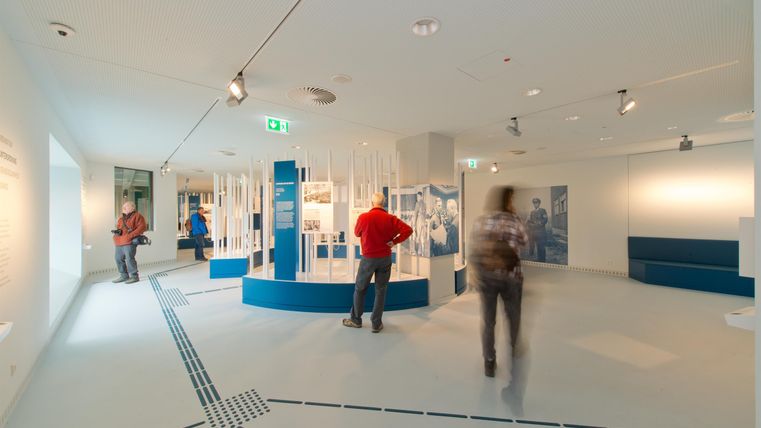 Ein modernes Ausstellungszimmer mit hellen Wänden und blauen Ausstellungsinseln. Besucher betrachten die Exponate und erkunden die Umgebung.