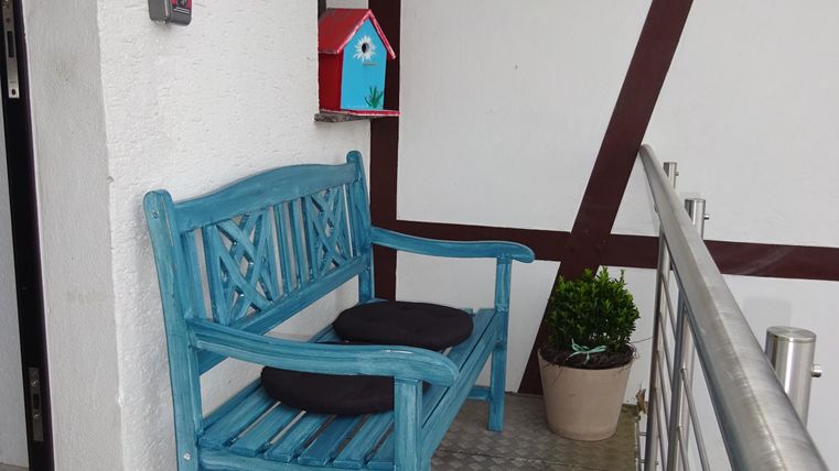 Eine blaue Bank steht auf einem Balkon, daneben ein kleiner Blumentopf. An der Wand hängt ein rotes Vogelhaus.