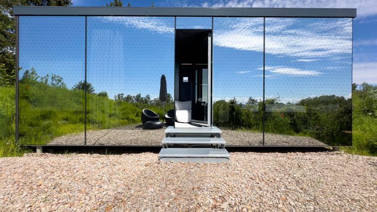 Ein modernes, spiegelt sichendes Gebäude in einer natürlichen Umgebung. Der Eingangsbereich ist von Kies umgeben und der Himmel ist blau mit einigen Wolken.