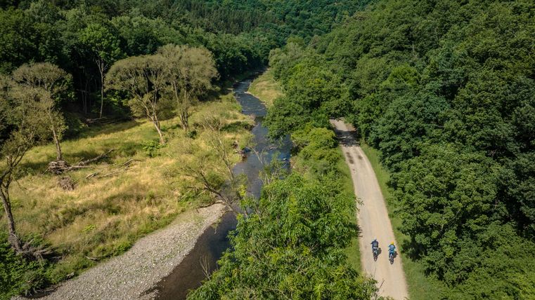 Zwei Radfahrer auf einem Weg neben einem Fluss in einer grünen Waldlandschaft.