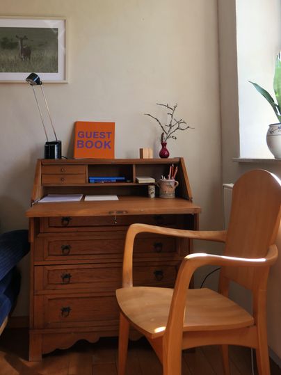 Ein Holzschreibtisch mit einer Schreibtischlampe und einem Gästebuch. Neben dem Schreibtisch steht ein Stuhl und eine Pflanze auf der Fensterbank.