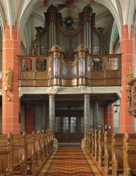 Eine große Orgel steht im oberen Teil einer Kirche. Der Innenraum ist mit Holzarbeiten und bunten Wänden dekoriert.