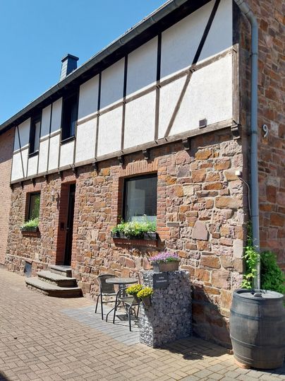 Ein charmantes Haus mit einem Steinverkleidung und einem Fachwerk-Obergeschoss. Vor dem Haus stehen ein Tisch und Stühle sowie ein Holzfass.