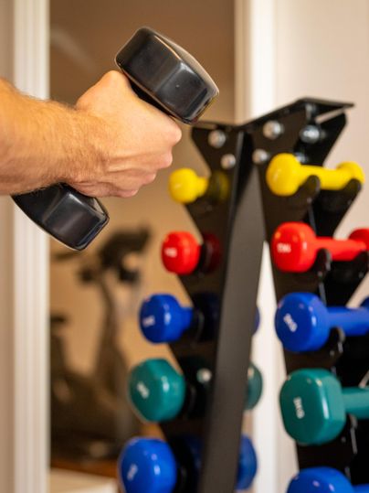 Eine Hand hält eine schwarze Hantel. Im Hintergrund steht ein Hantelständer mit bunten Gewichten.